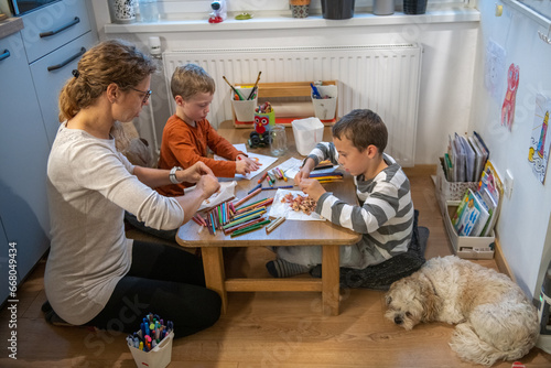 A mother and two children are drawing with crayons. they are sitting at the table. They are getting ready for school. They are sitting behind floor sitting desk