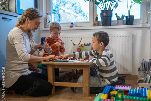 A mother and two children are drawing with crayons. they are sitting at the table. They are getting ready for school. They are sitting behind floor sitting desk