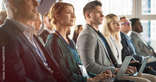 Young Woman Sitting in Crowded Audience at Business Conference. Female Delegate Smiling and Using Laptop Computer. Manager Watching Motivational Presentation About Investing In Innovative Startups.