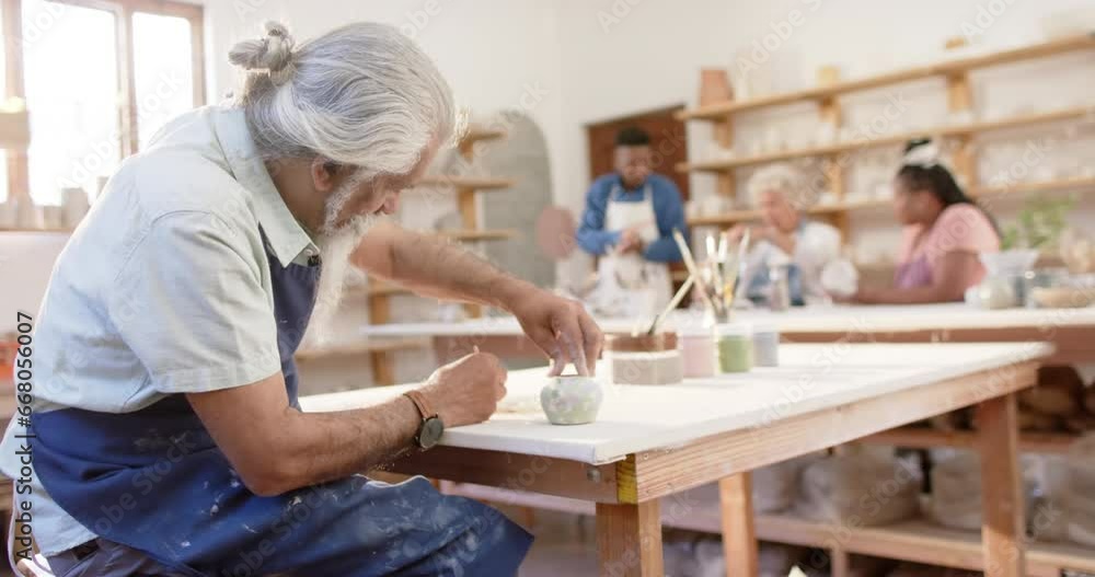 Focused biracial potter with long beard glazing clay jug in pottery studio, slow motion Focused biracial potter with long beard glazing clay jug in pottery studio, slow motion