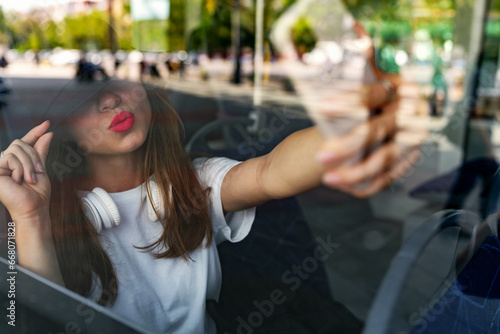 Stylish urban woman taking a selfie while sitting in city shuttle bus. City lifestyle, photography through the glass. Mobile phone messaging. photo