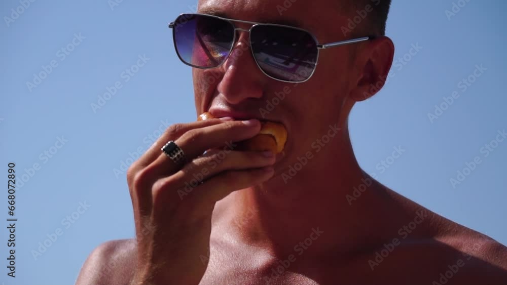 Young, happy man eats sandwich with cheese, sausage and ketchup on beach by sea. Takeaway food. Lunch break. Silhouette head of male tourist sitting on beach and eating sandwich, slow-motion. Profile Young, happy man eats sandwich with cheese, sausage and ketchup on beach by sea. Takeaway food. Lunch break. Silhouette head of male tourist sitting on beach and eating sandwich, slow-motion. Profile