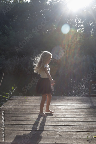 Adorable little blond girl dancing on a dock under sunlight. Idyllic summer day outdoor in nature
