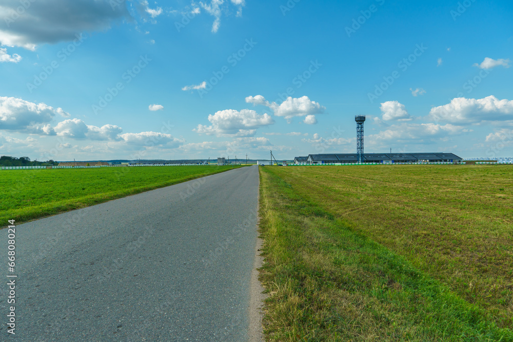 An empty roadway outside the city passes by agricultural fields. A highway with a new asphalt surface on the background of fluffy clouds. Paved road on a sunny day without cars. An empty roadway outside the city passes by agricultural fields. A highway with a new asphalt surface on the background of fluffy clouds. Paved road on a sunny day without cars.
