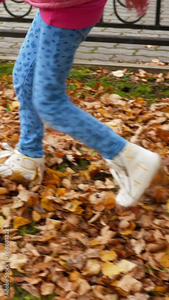 Slow motion of young anonymous girl in white boots walking on yellow dry leaves in autumn park, cropped view of child kicking foliage,vertical shot. Slow motion of young anonymous girl in white boots walking on yellow dry leaves in autumn park, cropped view of child kicking foliage,vertical shot.