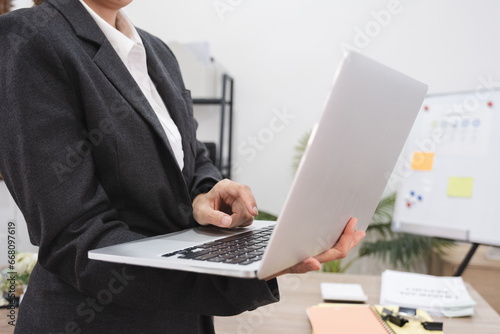 Close up, Young Asian businesswoman with a bright smile holds a laptop and stands in front of a work desk with a corporate planning board.