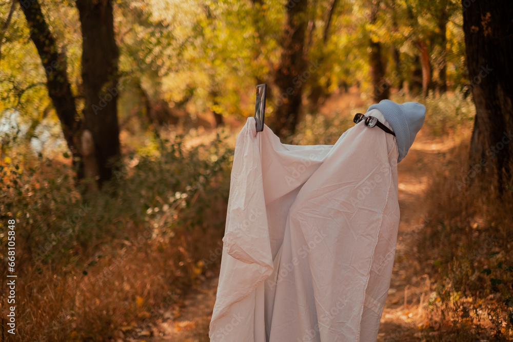A child in sheets and black glasses, holding a phone in his hands, looking like a ghost costume in the autumn forest, frightening and frightening. Nice little funny ghost. Halloween Party A child in sheets and black glasses, holding a phone in his hands, looking like a ghost costume in the autumn forest, frightening and frightening. Nice little funny ghost. Halloween Party
