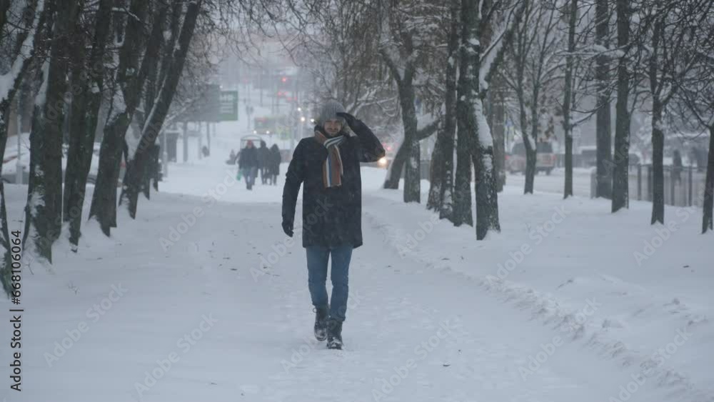 Man in warm clothes with scarf walks along sidewalk among trees and shields himself from heavy snowfall with his hand. Positive mood with smile during extreme winter weather in city. Man in warm clothes with scarf walks along sidewalk among trees and shields himself from heavy snowfall with his hand. Positive mood with smile during extreme winter weather in city.