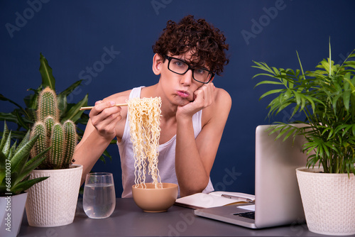 A funny young man in a white T-shirt eats noodles and teaches online lectures. A guy on a blue background with a laptop. Poor student.