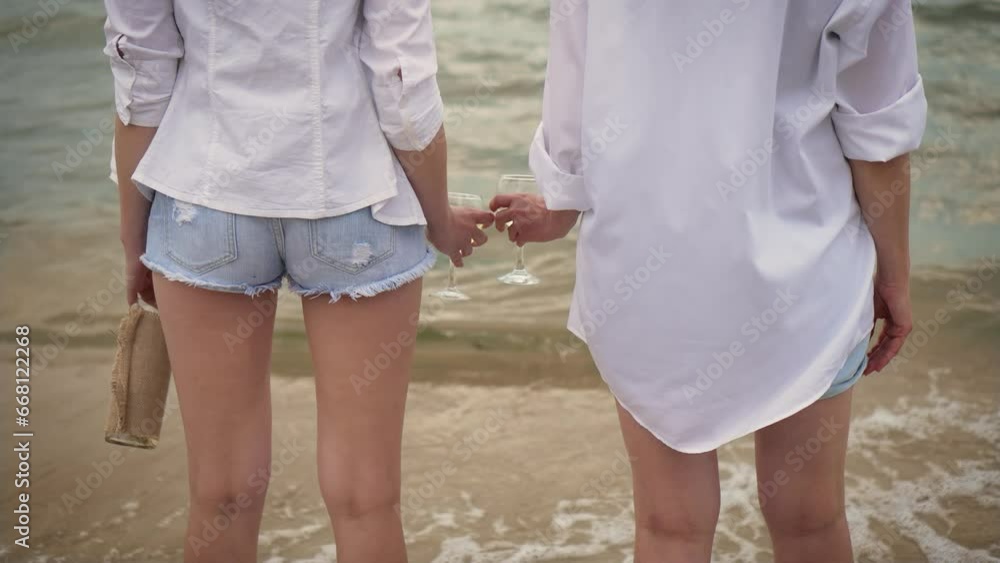 Unrecognizable females drinking wine at shoreline. Back view LGBT couple bonding together at beach. Lesbians bisexual transgender movements and love