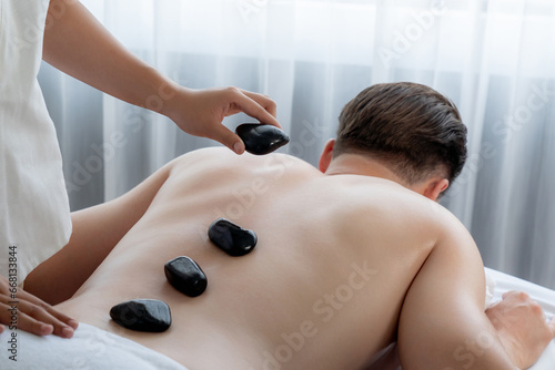 Hot stone massage at spa salon in luxury resort with day light serenity ambient, blissful man customer enjoying spa basalt stone massage glide over body with soothing warmth. Quiescent