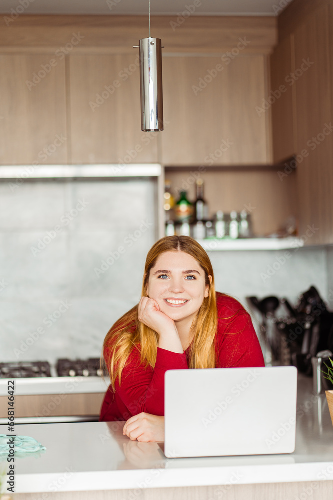 Portrait of smiling plus size woman using laptop, working at home in kitchen in cozy apartment Portrait of smiling plus size woman using laptop, working at home in kitchen in cozy apartment
