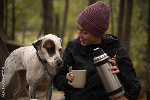 Girl in winter hat is sitting while drinking coffee with her dog outdoors.