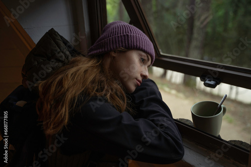 Girl in winter hat is looking out the window of a log cabin while drinking coffee from the cup.