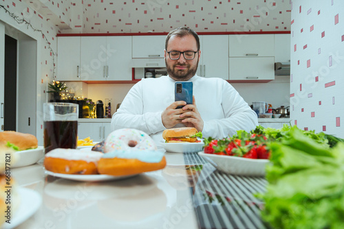 Man with extra weight photographing food on lunchtime at home in the kitchen. Concept of dieting and loosing weight, fatman only picturing food but not eat it