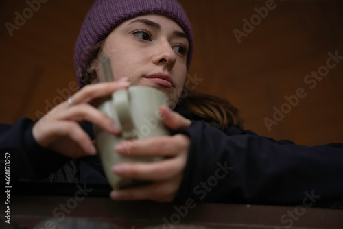 Girl in hat and autumn clothes is looking out the window of a log cabin while drinking coffee from the cup.