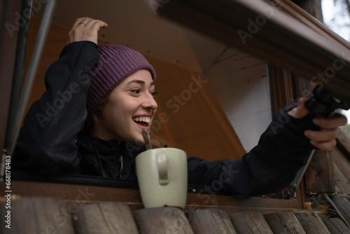 Girl in hat and autumn clothes is looking out the window of a cabin and smiles wooden smile while drinking coffee from the cup.