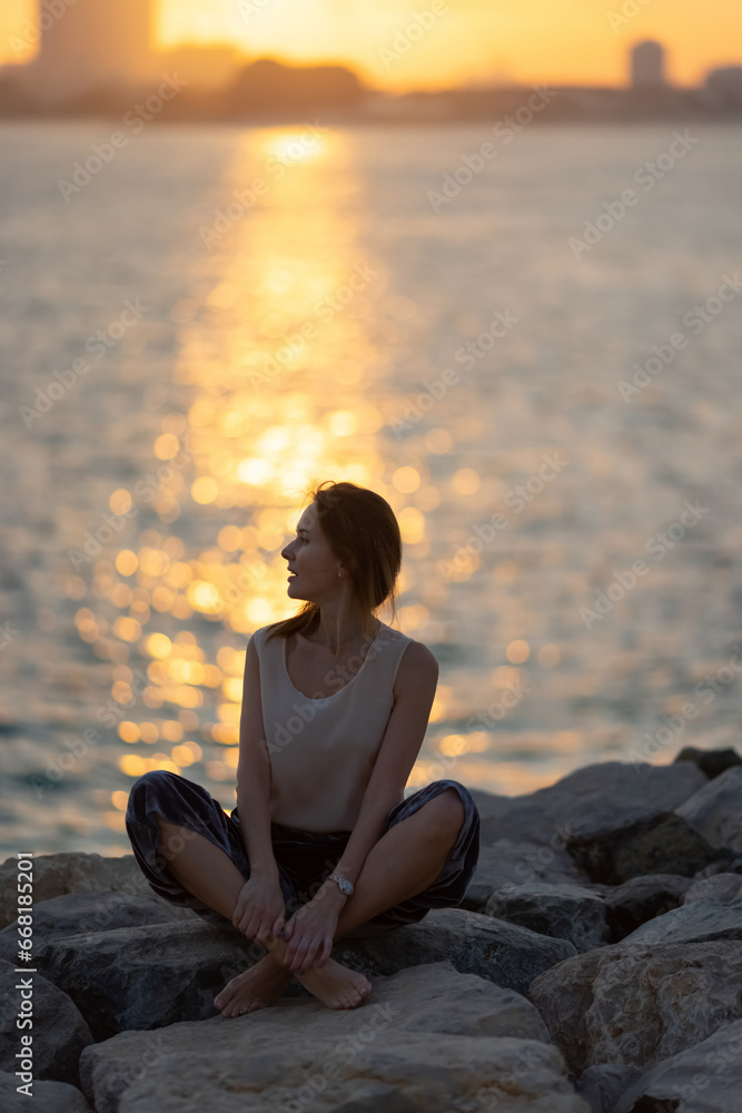 Woman practicing yoga meditation at sunrise. Morning yoga on the beach or coast of sea urban city background.