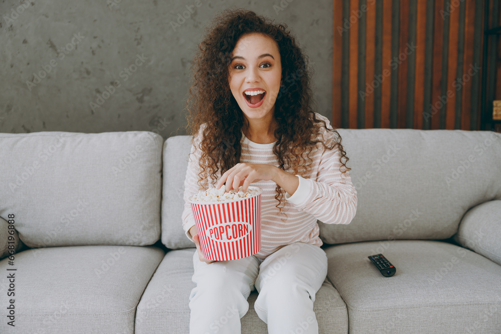 Φωτογραφία Young happy woman wear casual clothes sits on grey sofa couch eat popcorn watch movie film look camera stay at home hotel flat rest relax spend free spare time in living room indoor