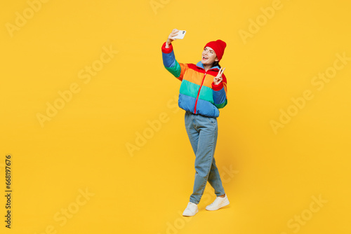 Full body side view young woman wears padded windbreaker jacket red hat casual clothes do selfie shot on mobile cell phone post photo on social network show v-sign isolated on plain yellow background.