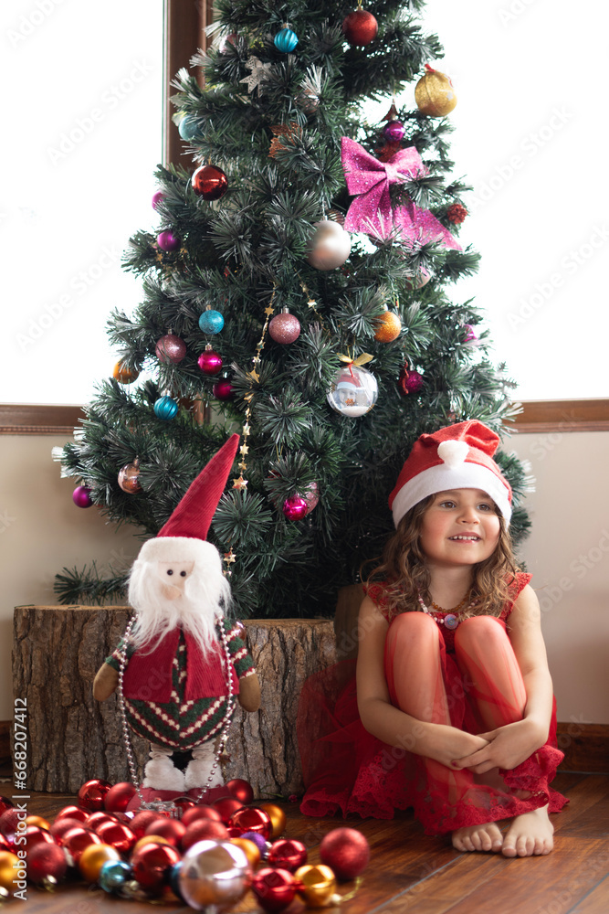 Little pretty girl in beautiful red dress playing near decorated Christmas tree with Xmas decoration and a fairy gnome. High quality photo