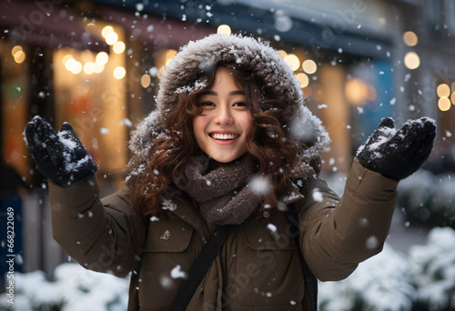 An attractive young Asian woman in dark brown furry winter coat standing outside catches snow with her hands on street with contented smile 