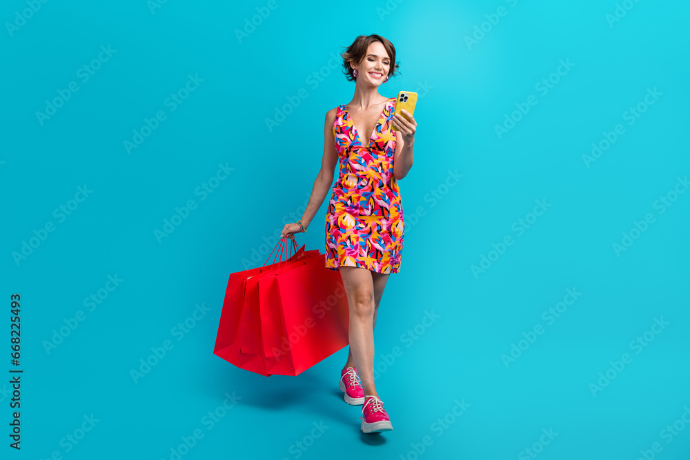 Full length photo of good mood girl wear colorful dress holding shopping bags look at smartphone isolated on blue color background Full length photo of good mood girl wear colorful dress holding shopping bags look at smartphone isolated on blue color background