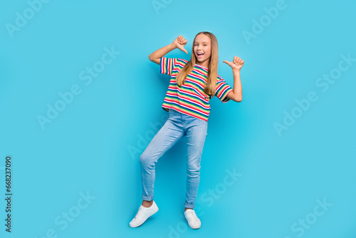 Full size photo of perky optimistic teen girl wear colorful t-shirt denim pants directing at herself isolated on blue color background