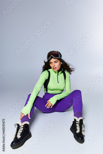 brunette sporty woman with curly hair posing in trendy active wear while sitting on grey backdrop