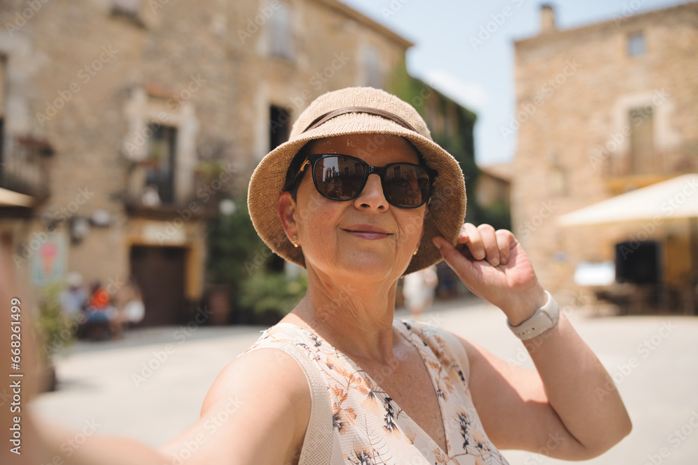 A retired woman in a straw hat and flower dress enjoys taking selfies walking around the picturesque village of Peratallada, Catalonia. A sunny day, in a Mediterranean heritage destination.