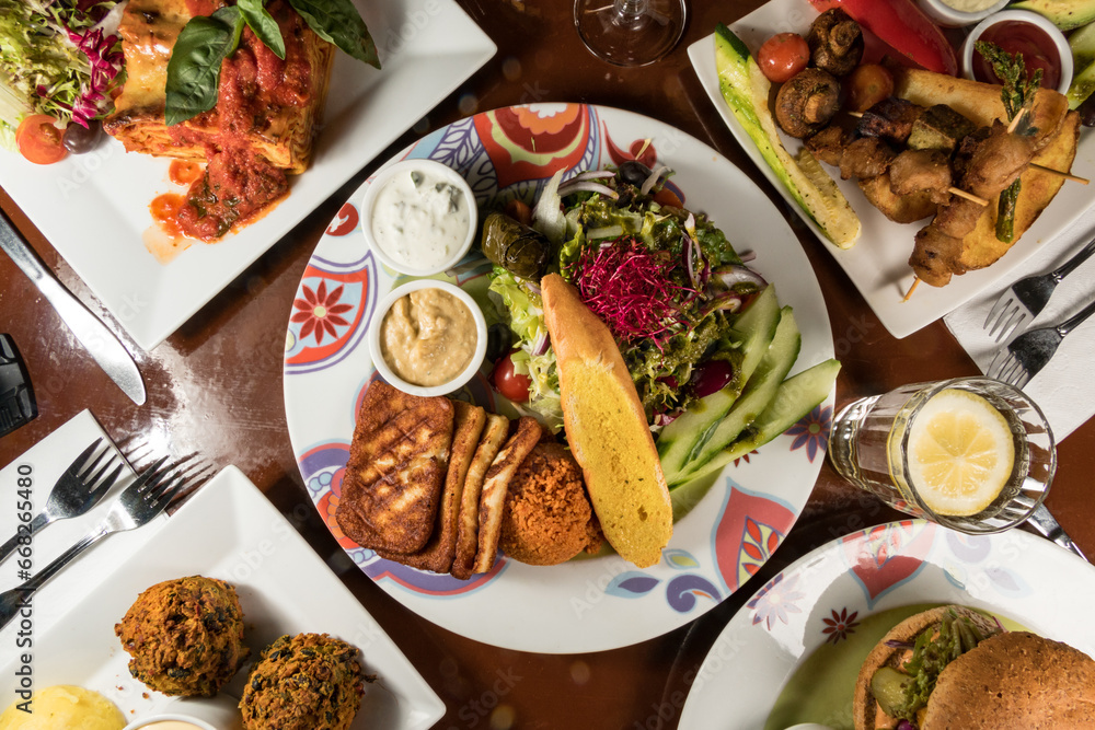 A plate of fried halloumi cheese with salad, bread and sauces surrounded by falafel, lasagne, and fried vegetable on a skewer.