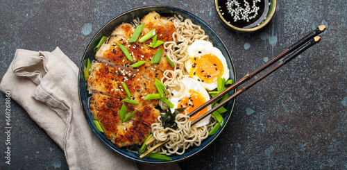 Asian noodles ramen soup with deep fried panko chicken fillet and boiled eggs in ceramic bowl with chop sticks and soy sauce on stone rustic background top view