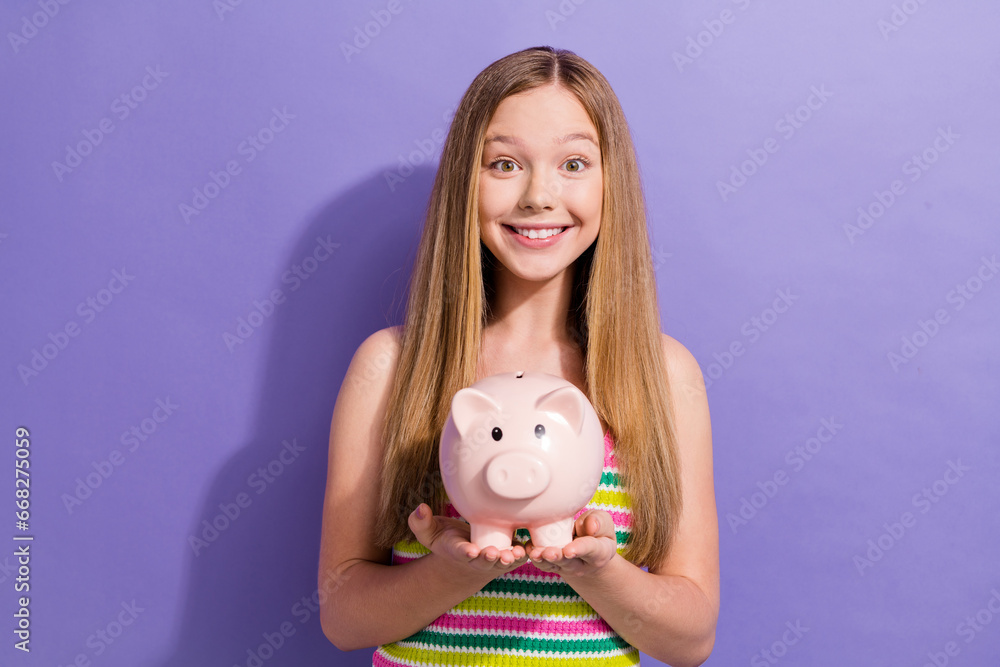 Photo of satisfied schoolgirl with straight hairdo dressed striped top hands holding piggy money box isolated on violet background Photo of satisfied schoolgirl with straight hairdo dressed striped top hands holding piggy money box isolated on violet background