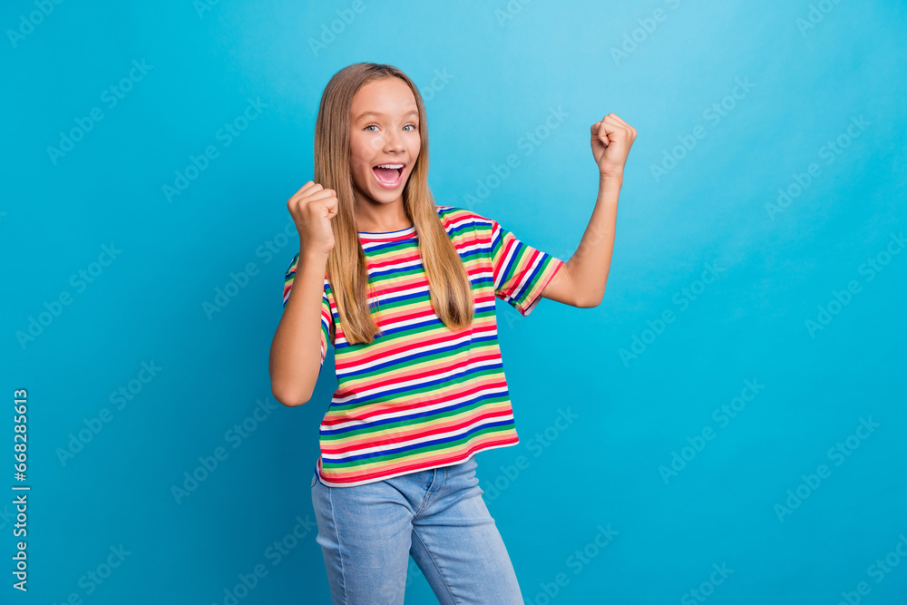 Portrait of funky overjoyed teen girl with long hair wear colorful t-shirt clenching fists pass exam isolated on blue color background Portrait of funky overjoyed teen girl with long hair wear colorful t-shirt clenching fists pass exam isolated on blue color background