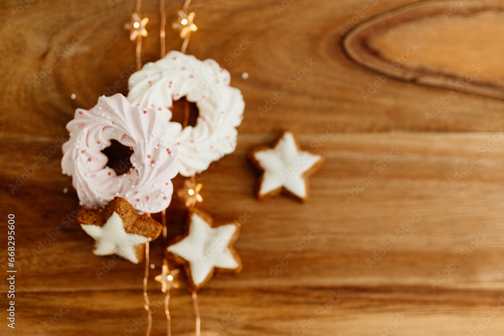 New Year background for winter cards. Gingerbread cookies in the shape of stars and pink meringue cookies lie on a brown background with Christmas lights. Delicious sweet pastries on a wood table