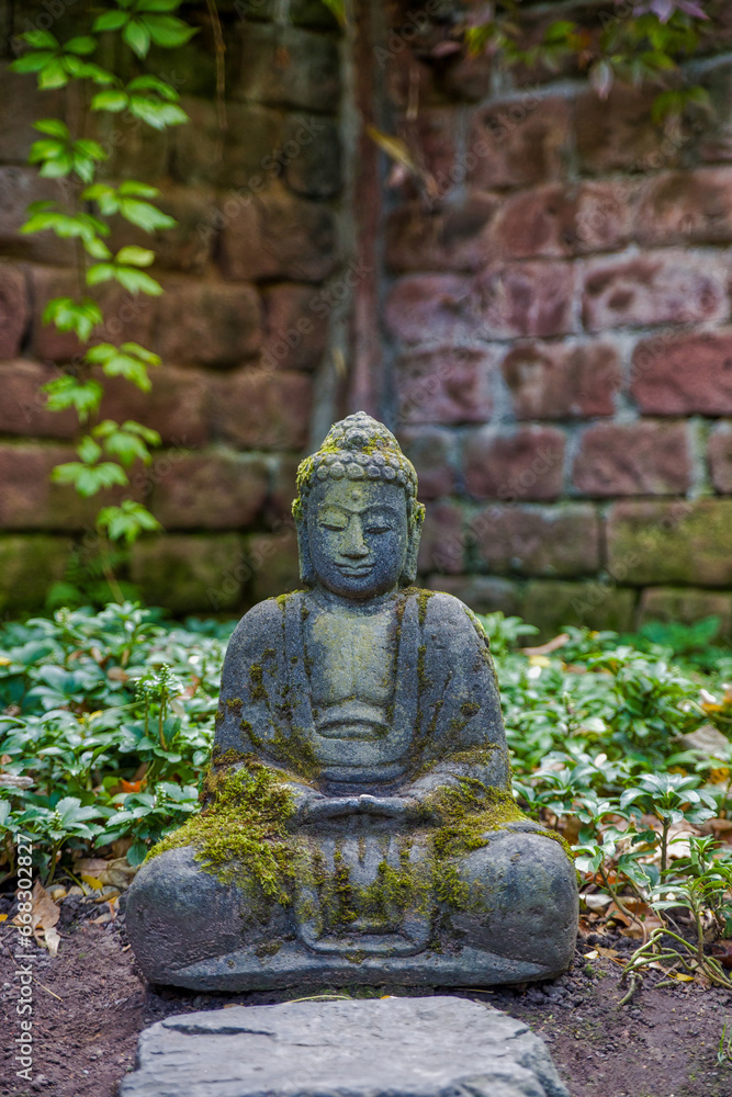 a grey stone buddha with moss in front of an old red brick wall and ivy