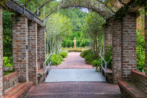 Fototapeta Naklejka Na Ścianę i Meble -  A long red brick footpath with a metal awning covered with lush green plants and trees with a gold statue at the end of the footpath at New Orleans Botanical Garden in New Orleans Louisiana USA