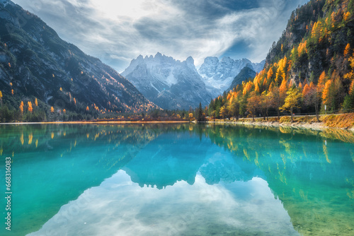 Fototapeta Naklejka Na Ścianę i Meble -  Mountain lake with reflection at sunny autumn day in Dolomites, Italy. Beautiful landscape with azure water, orange trees, snowy mountains, blue sky with clouds in fall. Snow covered rocks. Nature