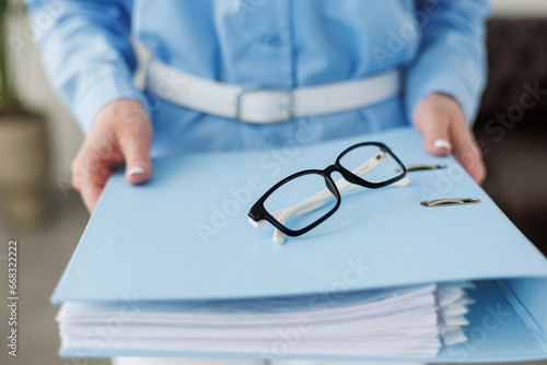 Cropped shot of unrecognisable business woman in blue formal shirt holding full folder of financial documents, selective focus on eyeglass, empty copy space place for advertising text message