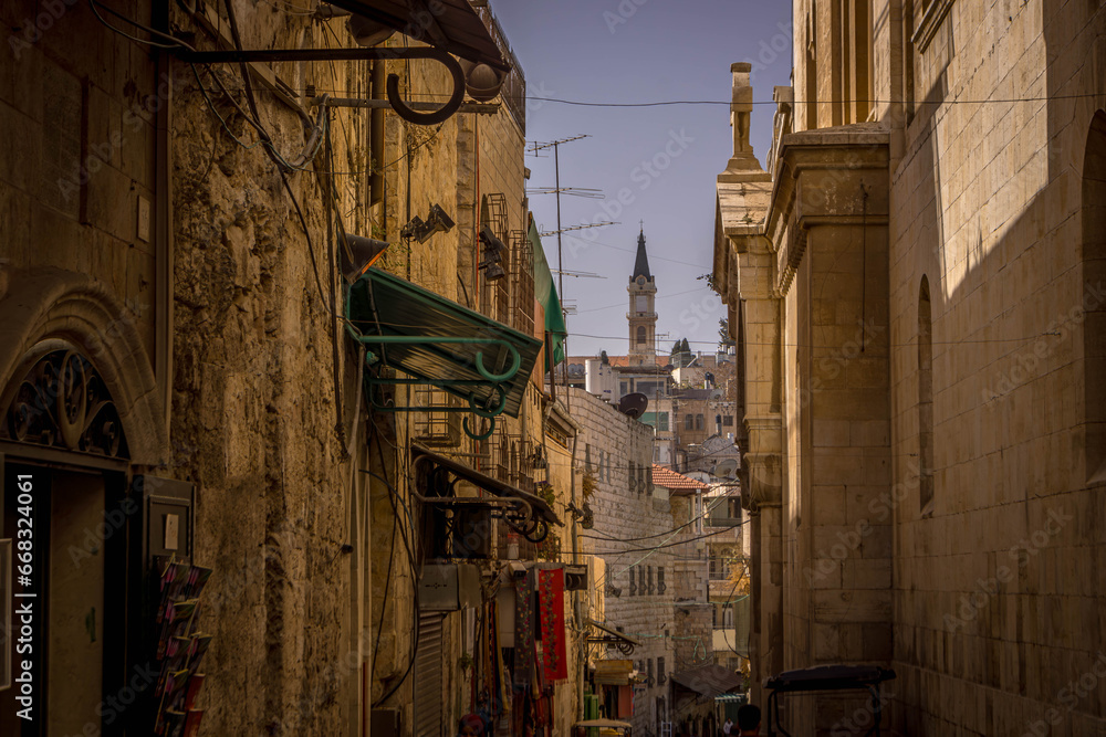 The tower of the Christian church is seen through the narrow street alley and historic buildings deep in Old Town of Jerusalem, Israel.
