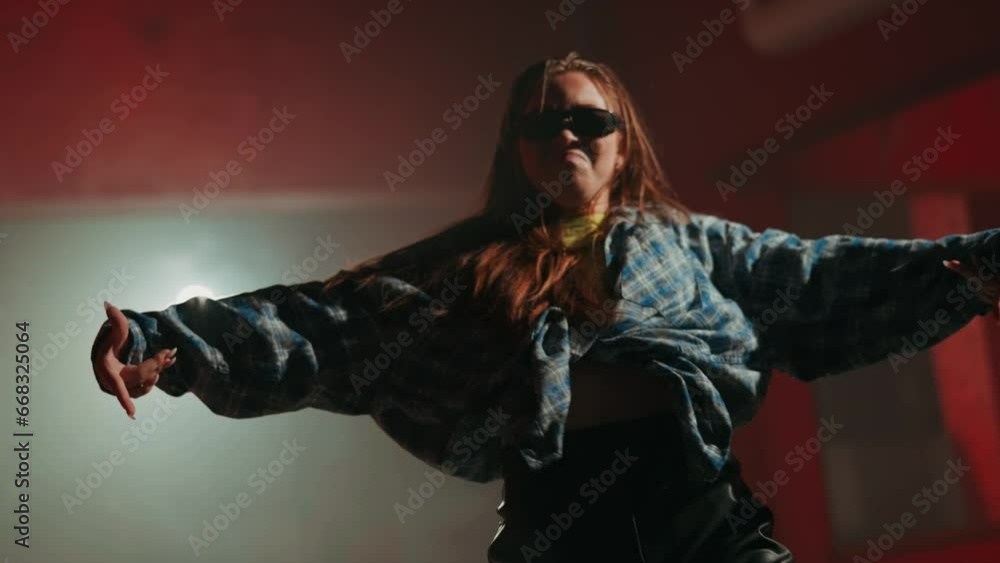 Young woman dancing hip hop in dance studio in white light background. Teenager wearing top with checkered shirt and black sunglasses doing dance freestyle movements closeup Young woman dancing hip hop in dance studio in white light background. Teenager wearing top with checkered shirt and black sunglasses doing dance freestyle movements closeup