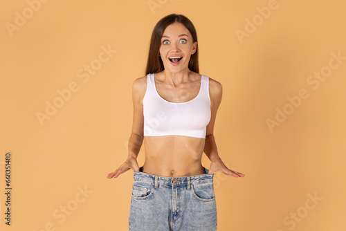 Slimming concept. Excited woman wearing big jeans after weight loss on peach studio background, smiling at camera with excitement, copy space
