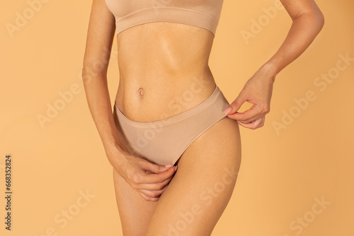 Fit woman posing in nude panties, showing hairless bikini line or smooth sexy hips without cellulite and stretch marks, peach background
