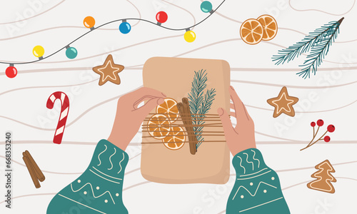 Process of packing a gift for Christmas. Female hands wrap a present in craft paper on wooden table. Organic decorations dry orange, gingerbread, spruce branch. Plastic free holiday concept