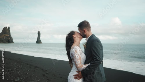 Bride and groom embracing and standing still in a long kiss. Iconic view of black sand on deserted beach, stormy sea with crashing waves, towering rocks. High quality 4k footage