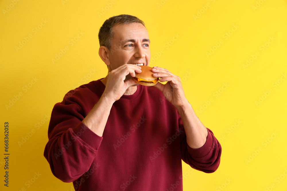 Mature man eating tasty burger on yellow background Mature man eating tasty burger on yellow background