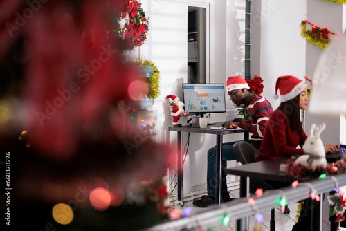 Focused african american businessman solving tasks on computer sitting at desk during holiday season. Meticulous employee wearing christmas hat, working in xmas decorated workplace office