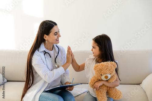 Doctor visit sick child at home. Pediatrician meets little patient during home visit, girl sits on coach, gives high five to doctor, medical care, shows warm attitude towards child concept. photo