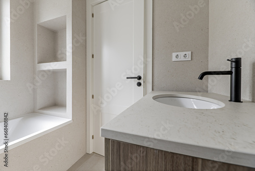 Image of the bathroom with cream-colored tiles  matching countertop  furniture with wooden drawers  black taps  white tub with built-in niche