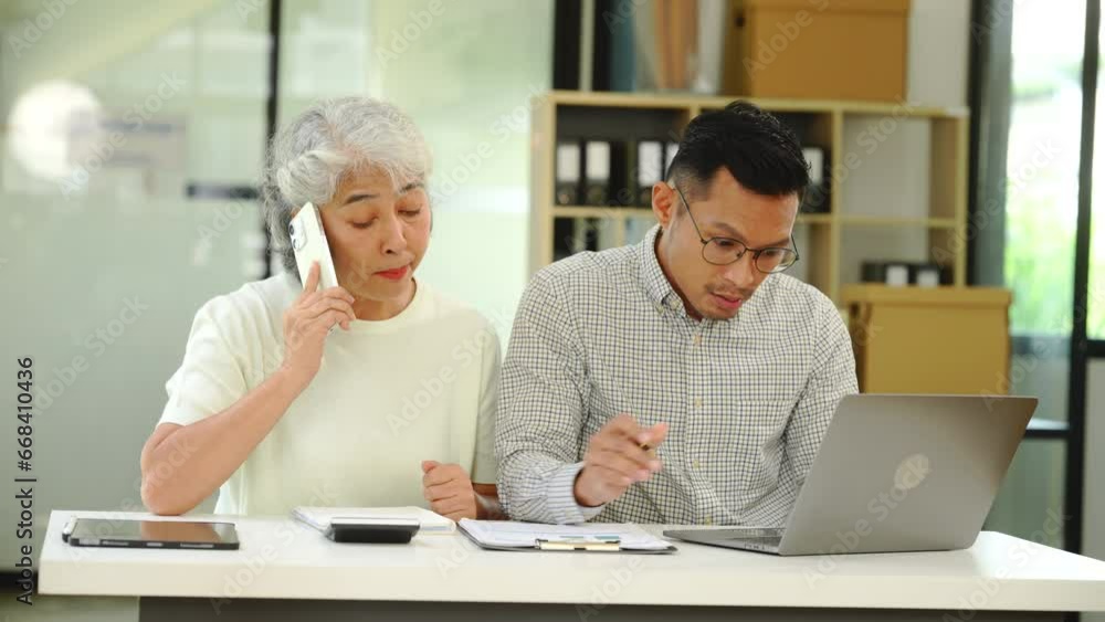 An older mature business woman and asian man work together. She has a tablet. He writes in a book. Both look at their notes. They are focused and happy. An older mature business woman and asian man work together. She has a tablet. He writes in a book. Both look at their notes. They are focused and happy.